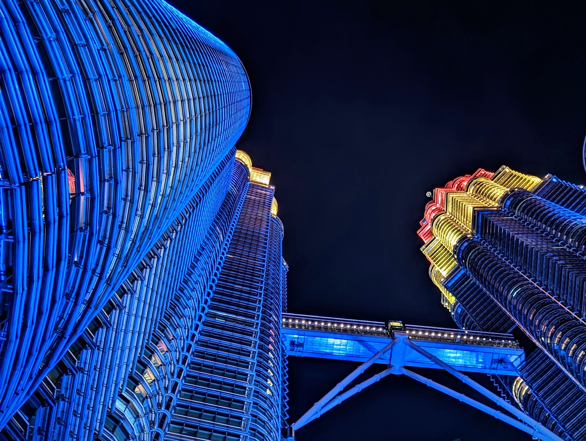 A photo of the Patronas Towers in KL from a steep angle below, with the towers lit in the blue, yellow, and red colors of the Malaysian flag.