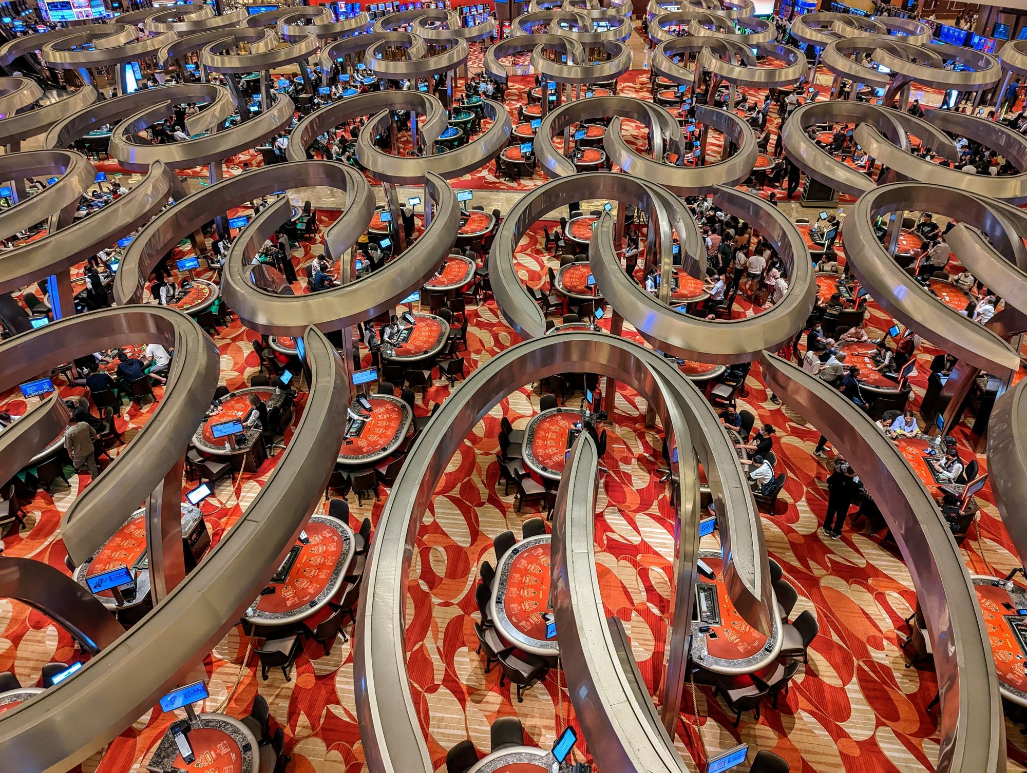 A casino floor seen from above, with striking golden lighting fixtures hanging over many gambling tables and a striking red carpet.